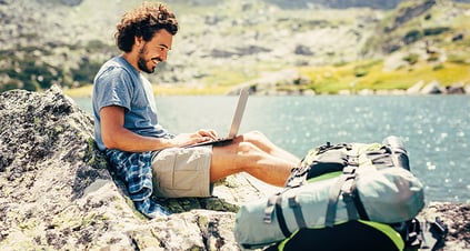 Man sitting on a rock next to a lake with a laptop on his lap